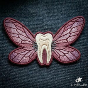 May include: An embroidered patch featuring a central off-white tooth shape flanked by two large, detailed insect wings in shades of mauve and dark purple. The wings have intricate vein patterns and a dark border. The text ElevenGifts is visible.
