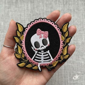 May include: An embroidered patch featuring a white skeleton with a pink bow on its head, centred within a black oval frame. The frame is bordered by a scalloped pink edge and decorated with gold embroidered leaves on the left and right sides.