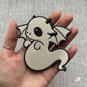 May include: An embroidered patch featuring a light beige dragon with a rounded body, small wings, and large black eyes. The design is outlined in thick black thread, creating a clear silhouette of a mythical creature with a curled tail.