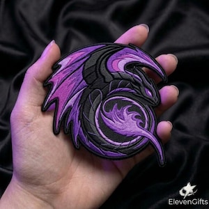 May include: A circular embroidered patch featuring a stylised dragon in shades of black and purple. The dragon is depicted in a coiled position with its wings spread, showcasing detailed stitching and a vibrant purple eye. The design is bordered by a thick black edge.