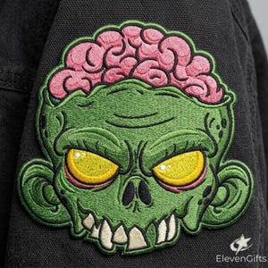 May include: An embroidered patch featuring a green zombie head with a prominent pink brain on top, yellow eyes, and white teeth. The patch is stitched onto a black fabric background. The text ElevenGifts is visible in the bottom right corner.