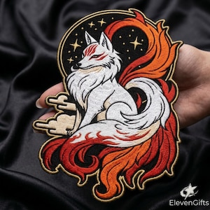 May include: An embroidered patch featuring a white multi-tailed fox with red facial markings, set against a black circular background with gold stars and clouds. The fox has a large, flowing tail with red and orange flame-like patterns.