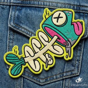 May include: An embroidered patch featuring a cartoon fish skeleton with a teal head, a pink tongue, and an X-shaped eye. The skeleton is cream-colored with a teal tail fin, all outlined in bright yellow thread against a blue denim fabric background.