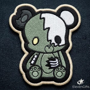 May include: An embroidered patch featuring a stylized teddy bear with a split design. One side is olive green with a yellow eye, while the other is white with a black eye socket. The bear has a torn chest revealing white stuffing and a small pencil.