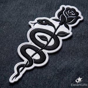 May include: An embroidered patch featuring a black snake coiled around a black rose with a white border. The design is rendered in black and white thread, showcasing the detailed texture of the snake scales and the petals of the rose flower.