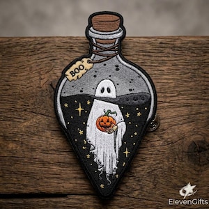 May include: An embroidered patch shaped like a glass bottle with a cork stopper. Inside the bottle is a white ghost holding a small orange pumpkin. The background is black with yellow stars. A small beige tag attached to the neck reads BOO.