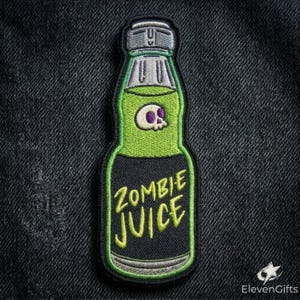 May include: An embroidered patch shaped like a bottle with a grey cap, a lime green upper section featuring a small white skull with purple eye sockets, and a black lower section with the yellow text ZOMBIE JUICE stitched in a bold font.