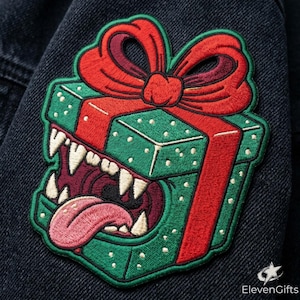 May include: An embroidered patch featuring a green gift box with a red ribbon and bow. The box has a wide-open mouth with sharp white teeth and a pink tongue protruding from the center. The design is stitched onto a dark blue denim fabric.