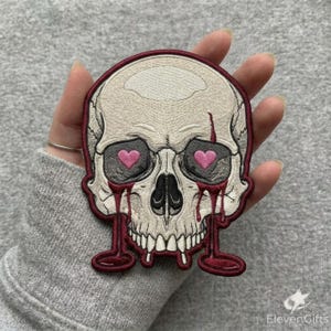May include: An embroidered patch featuring a cream-colored skull with dark outlines. The skull has pink heart-shaped pupils within its eye sockets and dark red liquid dripping from the eyes down to the bottom edge, framed by a dark red border.