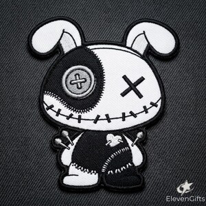 May include: An embroidered patch featuring a stylised rabbit character with a black and white colour scheme. The rabbit has one button eye, one X-shaped eye, stitched mouth details, and a heart patch on its chest with two pins inserted.