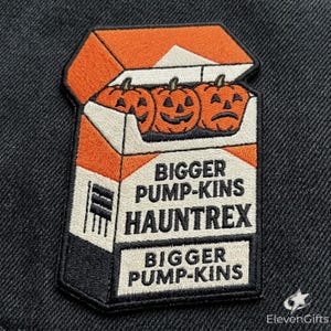 May include: An embroidered patch shaped like a cigarette pack with an orange and white design. It features three carved jack-o'-lantern pumpkins at the top and the text BIGGER PUMP-KINS HAUNTREX printed in black on the front of the pack.