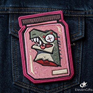 May include: An embroidered patch featuring a stylized green dinosaur head inside a pink jar. The dinosaur has a white eye with an X, sharp teeth, and a red tongue. The jar has a dark brown lid, pink liquid, and a small rectangular label at the bottom.