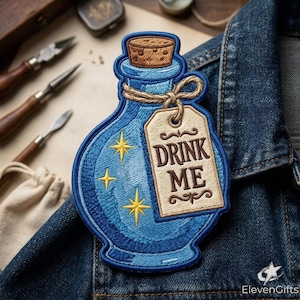 May include: An embroidered patch shaped like a blue potion bottle with a cork stopper. The bottle features yellow star accents and a beige rectangular tag tied with a string, displaying the brown text DRINK ME in a decorative font style.