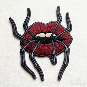 May include: An embroidered patch featuring a pair of dark red lips with white teeth and a black mouth interior. The lips are surrounded by six long, thin, dark grey spider-like legs extending outward from the center. The design has a black border.