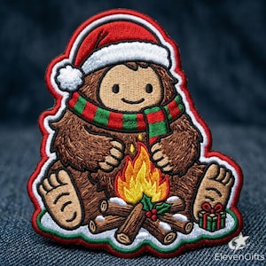 May include: An embroidered patch featuring a brown Bigfoot character wearing a red and white Santa hat and a red and green striped scarf. The character sits by a campfire with logs, flames, and a small green gift box. The text ElevenGifts is visible.