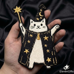 May include: An embroidered patch featuring a white cat dressed as a wizard in a black robe and pointed hat, both decorated with gold stars and a crescent moon. The cat holds a gold star-tipped wand in one paw and has a gold bell at the neck.