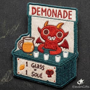 May include: An embroidered patch shaped like a lemonade stand with a red devil character, a yellow drink, and the text "DEMONADE" at the top and "1 GLASS = 1 SOUL" at the bottom, featuring a small lemon icon and a skull icon on the front panel.