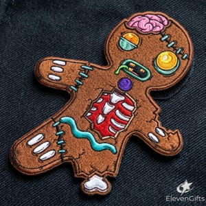 May include: An embroidered patch shaped like a brown gingerbread person with a zombie theme. It features a pink exposed brain, a red ribcage, a green tongue, and blue stitches. The design includes yellow and orange eyes and a purple circular button.