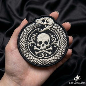 May include: A circular embroidered patch featuring a white snake biting its own tail in an ouroboros design. Inside the snake circle is a white skull and crossbones motif on a black background with decorative scrollwork accents.