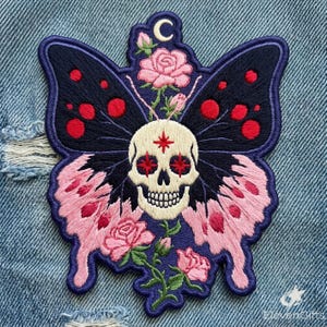 May include: An embroidered patch featuring a central cream-colored skull with red star-shaped eyes and a red star on the forehead. The skull is flanked by butterfly wings in dark blue with red spots and light pink with dark pink spots, accented by pink roses.