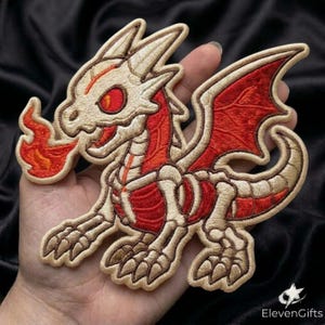 May include: An embroidered patch featuring a skeletal dragon design with cream-colored bones and red accents. The dragon is depicted in a side profile, breathing a flame from its mouth, with detailed stitching defining its wings and claws.