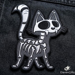 May include: An embroidered patch featuring a stylized black cat with a white skeletal rib cage, spine, and skull head. The cat has large dark eye sockets, two sharp fangs, and a long, curved, striped tail. The design is rendered in black and white.