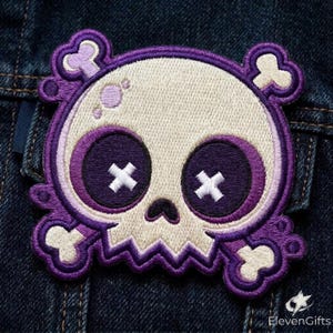 May include: An embroidered patch featuring a stylized skull with a cream-colored cranium, deep purple eye sockets, and white cross-shaped pupils. The skull is framed by a purple border with four small bones extending from the corners.