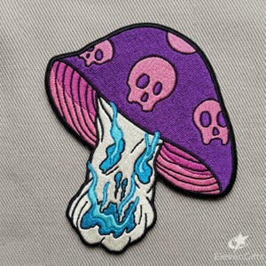 May include: An embroidered patch featuring a purple mushroom cap with pink skull patterns and pink gills. The mushroom stem is white with blue liquid-like details dripping down. The text ElevenGifts is visible in the bottom right corner.