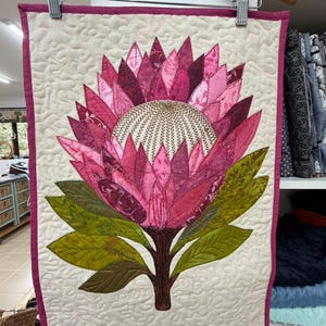 K&ouml;nnte beinhalten: Ein rechteckiger Wandbehang aus Stoff mit einer gro&szlig;en Protea-Bl&uuml;tenapplikation in der Mitte. Die Bl&uuml;te hat geschichtete rosa und magentafarbene Bl&uuml;tenbl&auml;tter mit einer strukturierten, cremefarbenen Mitte. Gr&uuml;ne Bl&auml;tter ragen vom Stiel ab.