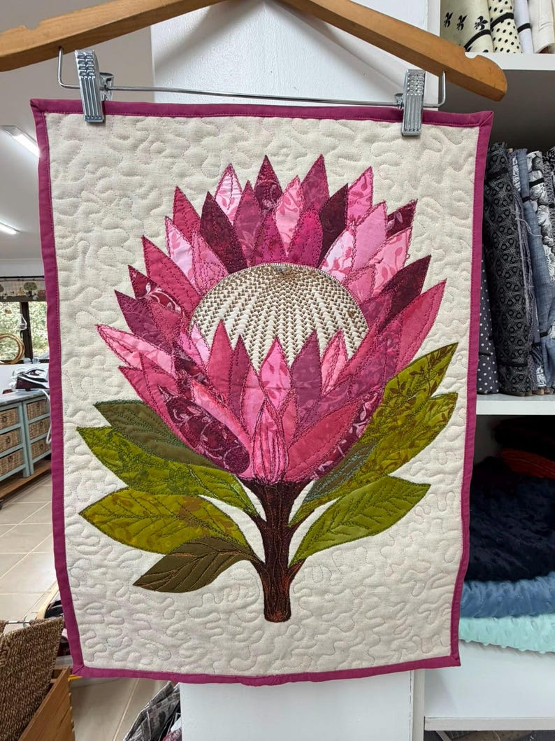 K&ouml;nnte beinhalten: Ein rechteckiger Wandbehang aus Stoff mit einer gro&szlig;en Protea-Bl&uuml;tenapplikation in der Mitte. Die Bl&uuml;te hat geschichtete rosa und magentafarbene Bl&uuml;tenbl&auml;tter mit einer strukturierten, cremefarbenen Mitte. Gr&uuml;ne Bl&auml;tter ragen vom Stiel ab.