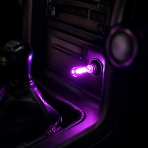 May include: A small, cylindrical purple LED light plug inserted into a vehicle's 12-volt accessory power outlet. The device emits a bright, vibrant purple glow that illuminates the surrounding black plastic center console and gear shift area.