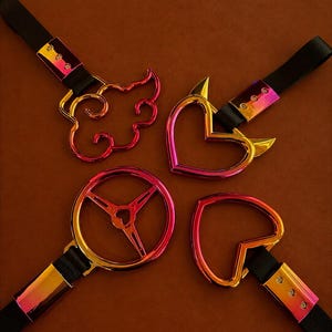 Peut inclure: Quatre poignées en métal irisé avec des sangles en nylon noir. Les formes incluent un nuage, un cœur avec des cornes de diable, un design de volant circulaire et un cœur simple. Chaque poignée présente une finition arc-en-ciel et trois rivets.