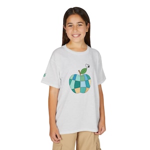 Puede incluir: Una camiseta de manga corta gris claro con un gráfico central de una manzana diseñada con un patrón de patchwork en tonos verde azulado, azul y amarillo. La manzana tiene un tallo marrón, una hoja verde y un pequeño corazón negro.