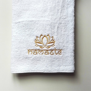 Puede incluir: Toalla de baño blanca con bordado dorado de una flor de loto y la palabra "namaste".
