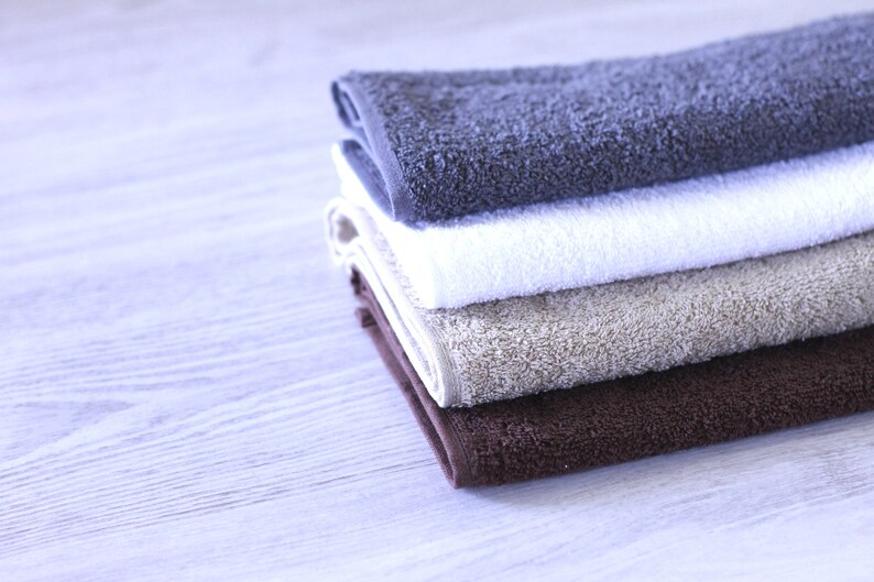 May include: A stack of four towels in different colors and textures. The top towel is dark gray, the second is white, the third is beige, and the bottom towel is brown.