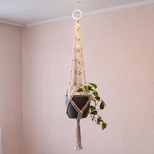May include: A cream-colored macrame plant hanger featuring intricate knotted patterns and a circular top ring. The hanger holds a dark gray pot containing a green leafy plant, with a long decorative tassel hanging from the bottom center.