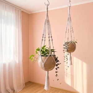 Pode incluir: Dois suportes de plantas em macramé branco suspensos por ganchos no teto. Cada suporte apresenta padrões de nós intrincados e uma borla longa na parte inferior, segurando um vaso de cerâmica redondo e liso em tom castanho claro.