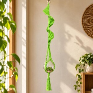 Può includere: Un portapiante in macramè verde lime brillante con un design a corda intrecciata a spirale sospeso a un anello di legno. Il supporto sostiene un terrario in vetro sferico trasparente contenente una piccola pianta grassa e termina con una lunga nappa.