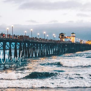 Könnte beinhalten: Ein hölzerner Steg erstreckt sich über ein blaues Meer mit weißen Wellen, die an das Ufer schlagen. Der Steg ist mit Lichterketten beleuchtet und hat ein kleines Gebäude am Ende. Der Himmel ist hellblau mit weißen Wolken. Der Text "Luxe Visuals Photography" ist in weiß unten links im Bild geschrieben.