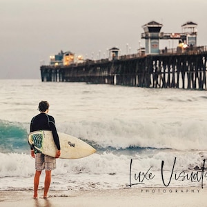 Könnte beinhalten: Ein Surfer mit einem Surfbrett steht am Strand und blickt auf das Meer. Im Hintergrund befindet sich ein Pier. Der Text "Luxe Visuals Photography" befindet sich unten rechts im Bild.