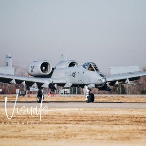 May include: A gray A-10 Thunderbolt II aircraft with its landing gear down, taxiing on a runway. The aircraft has a large, powerful engine on the left side and a smaller engine on the right side. The aircraft is in the process of landing.
