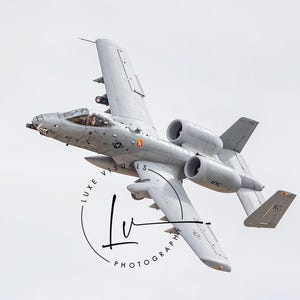 May include: A gray A-10 Thunderbolt II aircraft in flight. The aircraft is in a slight bank, with the wings tilted upwards. The aircraft has a single pilot in the cockpit, and the cockpit canopy is open. The aircraft is flying against a white background.