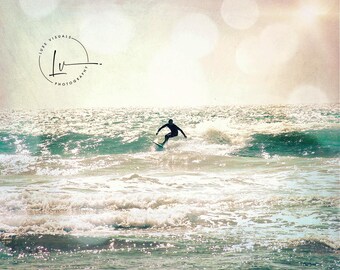 May include: A silhouette of a surfer riding a wave in the ocean. The water is a turquoise blue and the sky is a light blue with a soft, faded look. The image has a vintage feel.