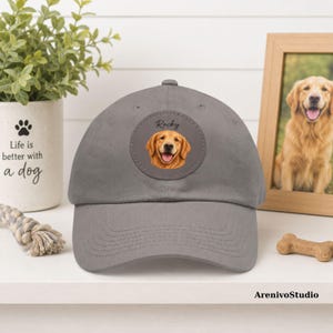 May include: A grey baseball cap featuring a circular patch on the front panel. The patch displays a realistic illustration of a golden retriever dog with its tongue out, positioned below the handwritten-style text Rocky in black ink.