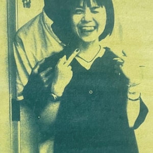 May include: A monochromatic yellow-toned print featuring two individuals. One individual stands behind the other, who is smiling with their tongue out and gesturing with a middle finger. The print has a grainy, high-contrast texture throughout.