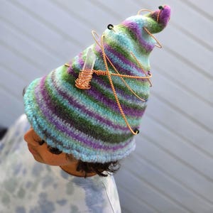 May include: A pointed hat featuring horizontal stripes in shades of purple, green, and light blue. The hat is adorned with a clear, elongated crystal wrapped in copper wire, with additional copper wire spiraling around the exterior of the hat.