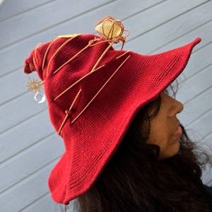 May include: A deep red knitted hat with a wide brim and a pointed crown. The hat is wrapped in thin copper-colored wire, featuring a large yellow crystal stone at the top and a small gold-toned sun charm with a clear crystal bead hanging from the side.