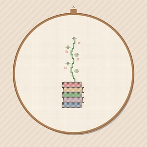 May include: A cross-stitch embroidery design inside a circular wooden hoop featuring a stack of five books in red, orange, green, purple, and blue. A single green vine with small green and pink cross-stitched flower accents grows upward.