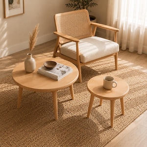 May include: A light wood armchair with a woven cane backrest and a cream-colored seat cushion, accompanied by two round light wood coffee tables of different sizes, a ceramic vase with dried grass, a small bowl, and a book on the larger table.