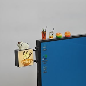Puede incluir: Una caja de luz negra montada en el lateral de un monitor con un perro de dibujos animados en una taza de café y el texto Coffee. Una figura de gato blanco se asienta encima. Dos vasos miniatura y dos figuras de fruta naranja están arriba.